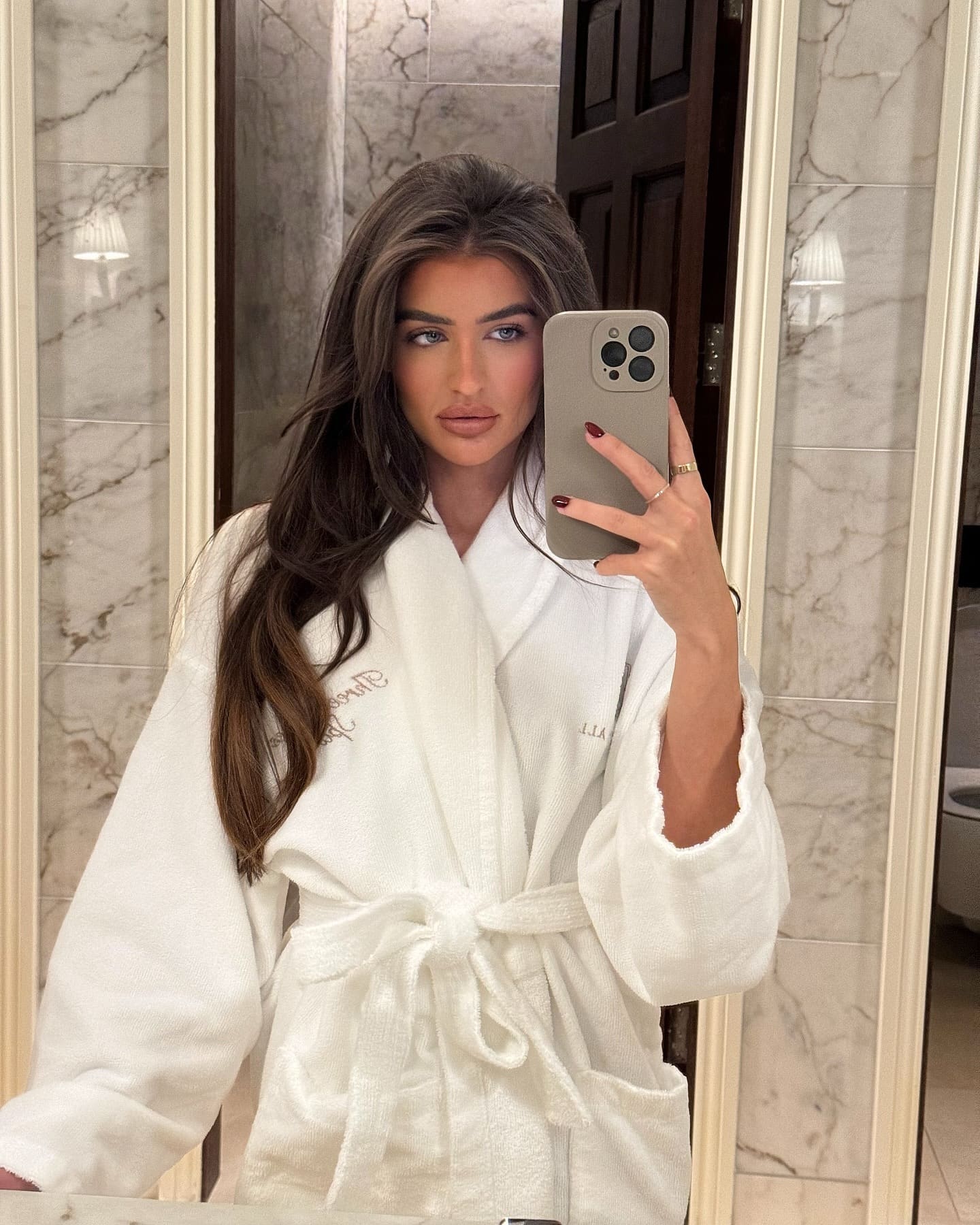 Alisa stands in a marble-tiled bathroom, wearing a white waffle-knit bathrobe and holding a gray smartphone for a mirror selfie.