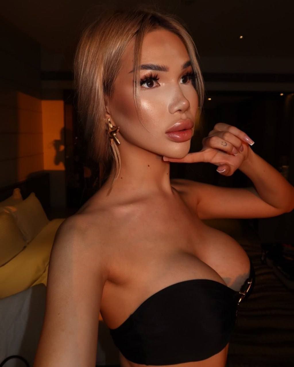 A woman in a black strapless top and gold earrings poses near a bed in a dimly lit room.