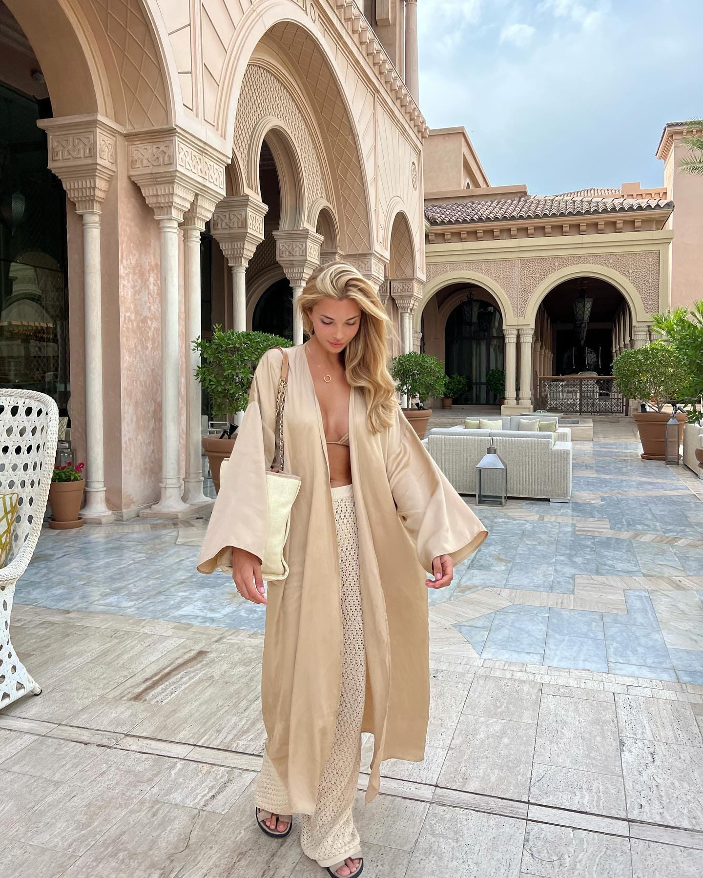 A blonde woman stands on a stone patio wearing a beige robe over a bikini top and patterned pants, carrying a small shoulder bag.