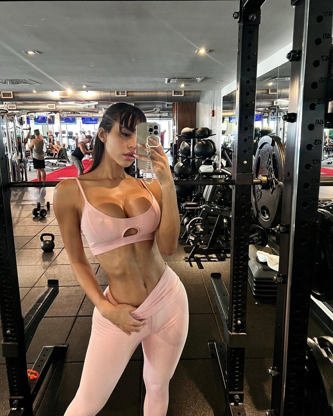 A woman takes a mirror selfie in a gym, wearing light pink sports bra and leggings, with weight racks and dumbbells visible in the background.