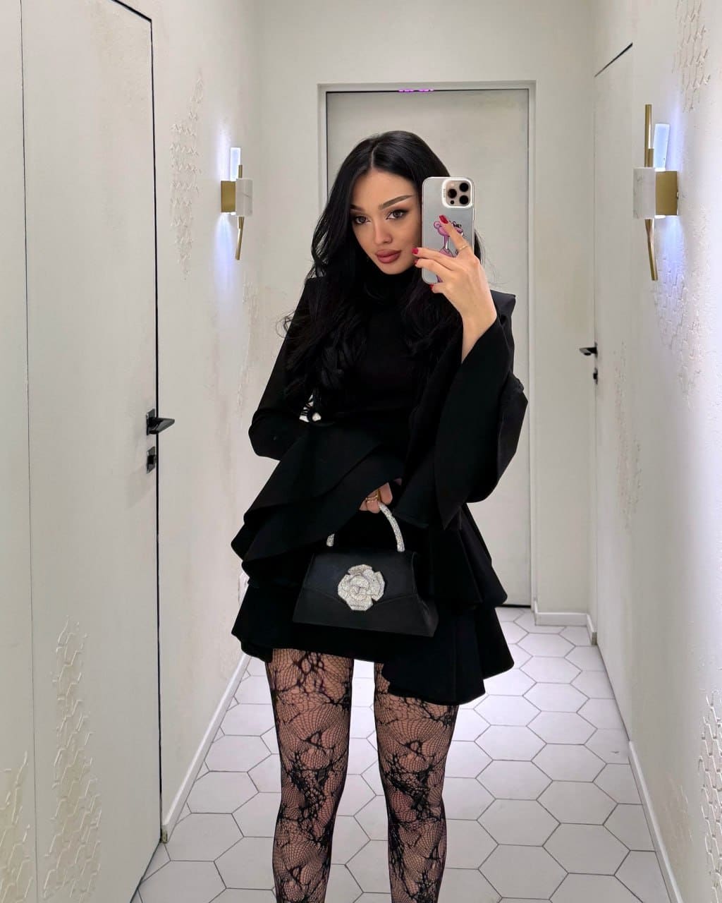 Mirror selfie in a white-tiled hallway showing a woman in a black ruffled dress, lace tights, and floral handbag.