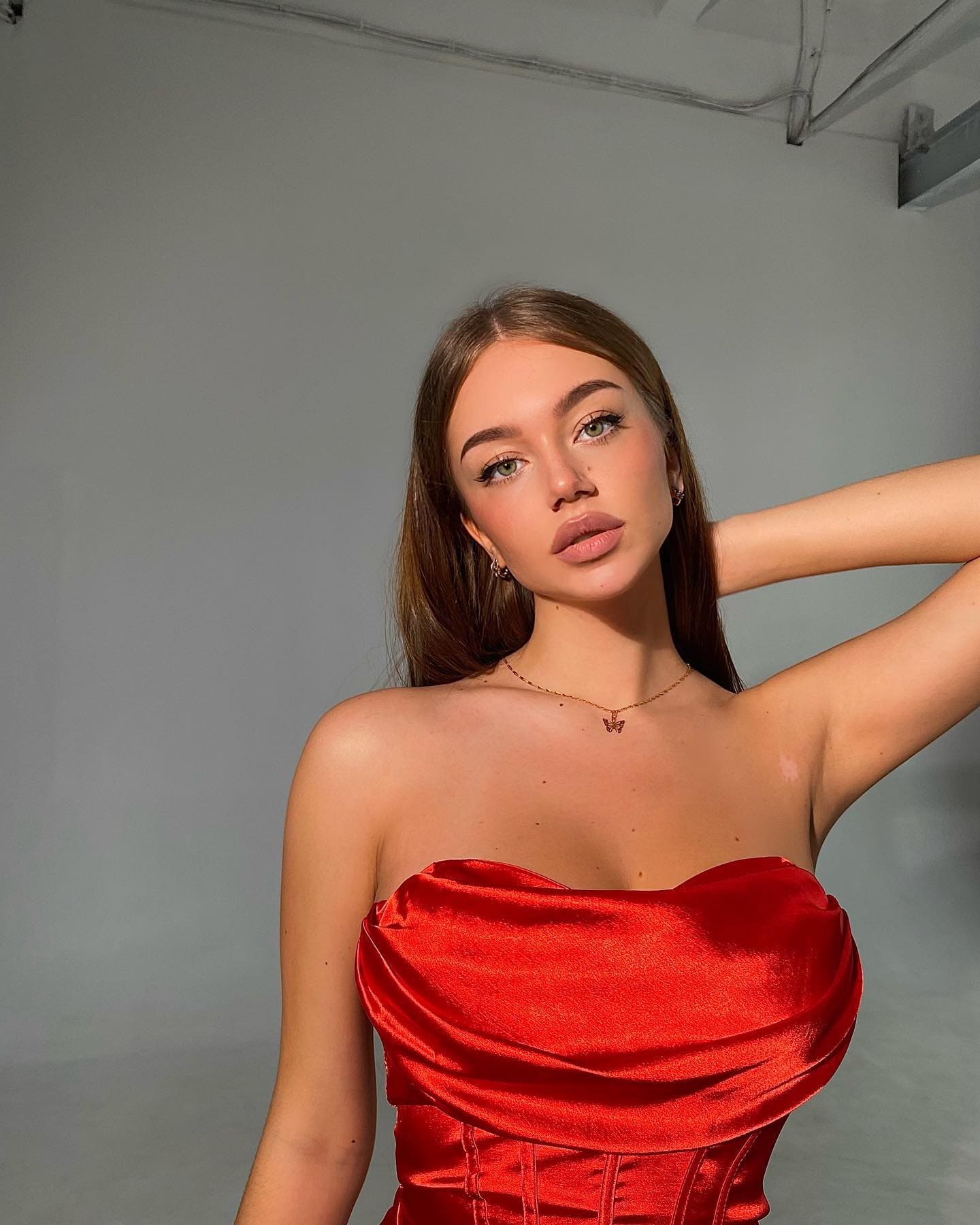 Inga wears a red strapless satin dress, posing with an arm raised against a gray studio wall.