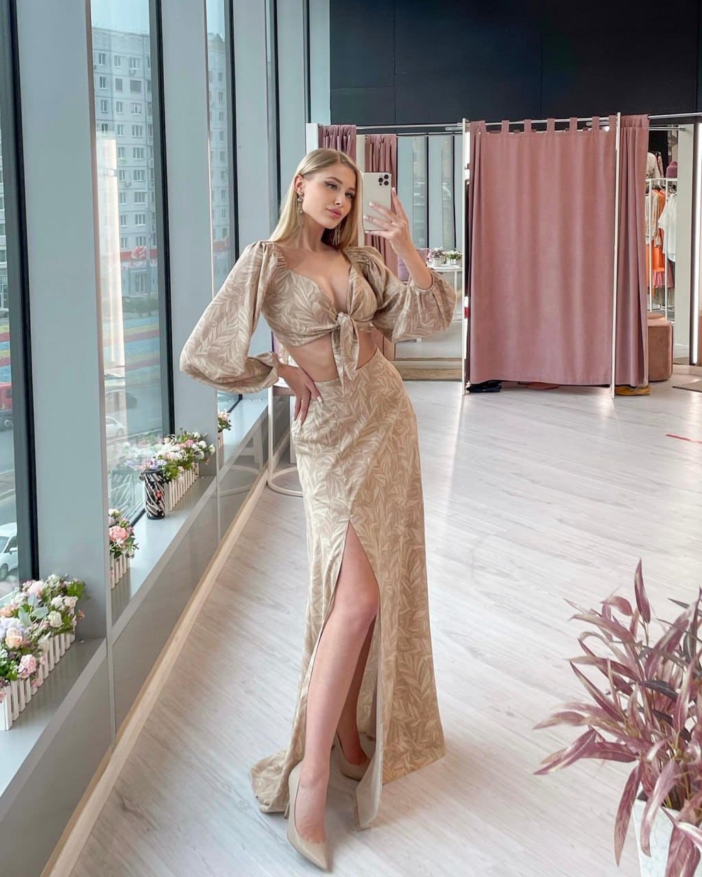 A blonde woman poses in a mirror selfie wearing a beige patterned two-piece outfit with a high-slit skirt in a bright room.