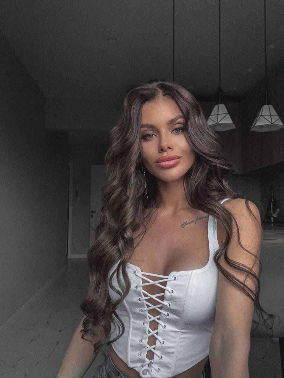 Daria stands indoors in a dimly lit room with pendant lights, wearing a white lace-up corset top with a deep square neckline.
