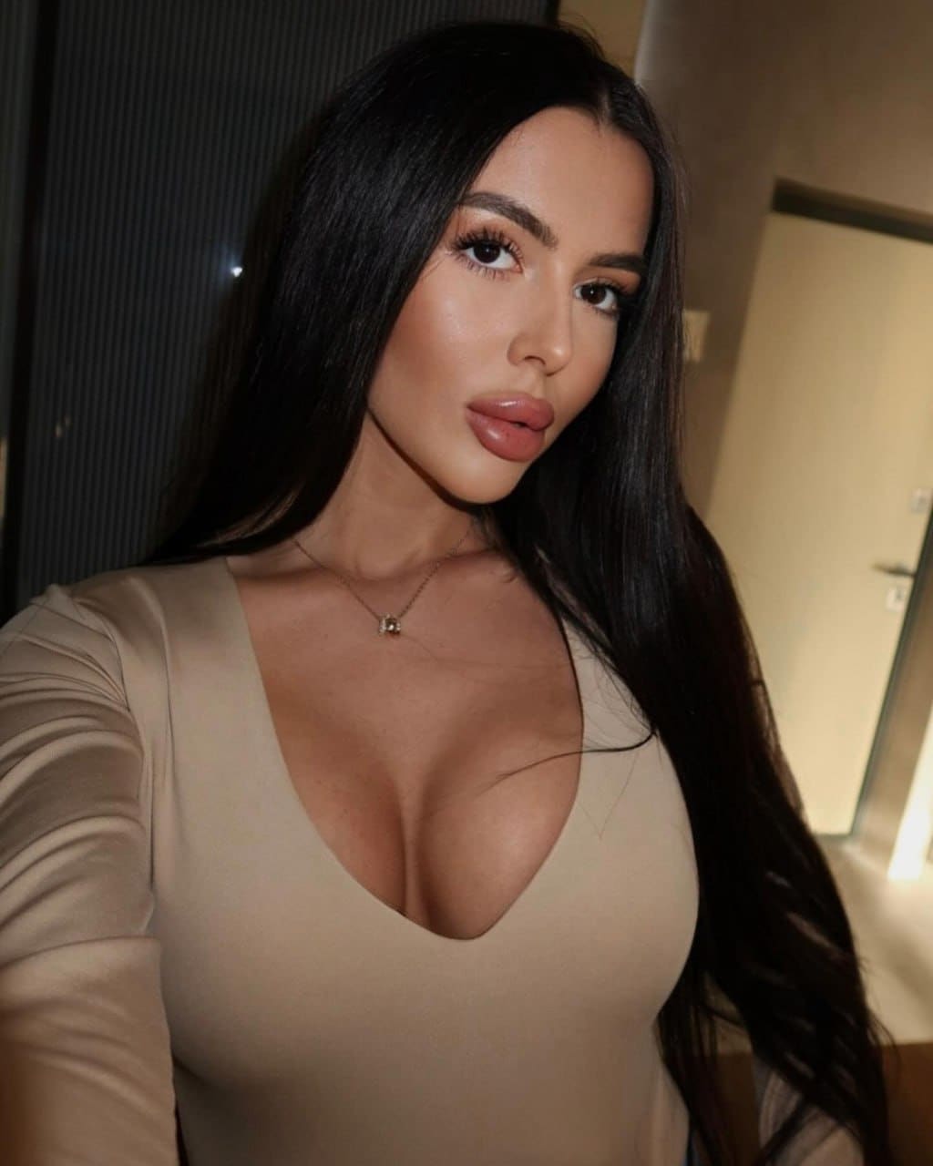 A woman takes a selfie in a beige long-sleeved top with a deep V-neck, with a door and dark wall visible behind her.