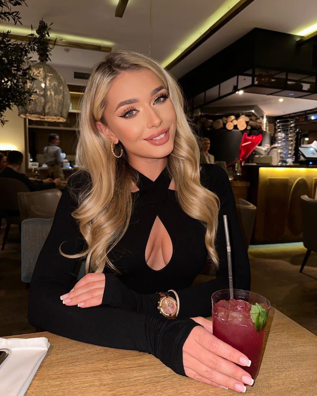 Sabrina sits at a wooden restaurant table in a black keyhole top, holding a red drink with a straw.