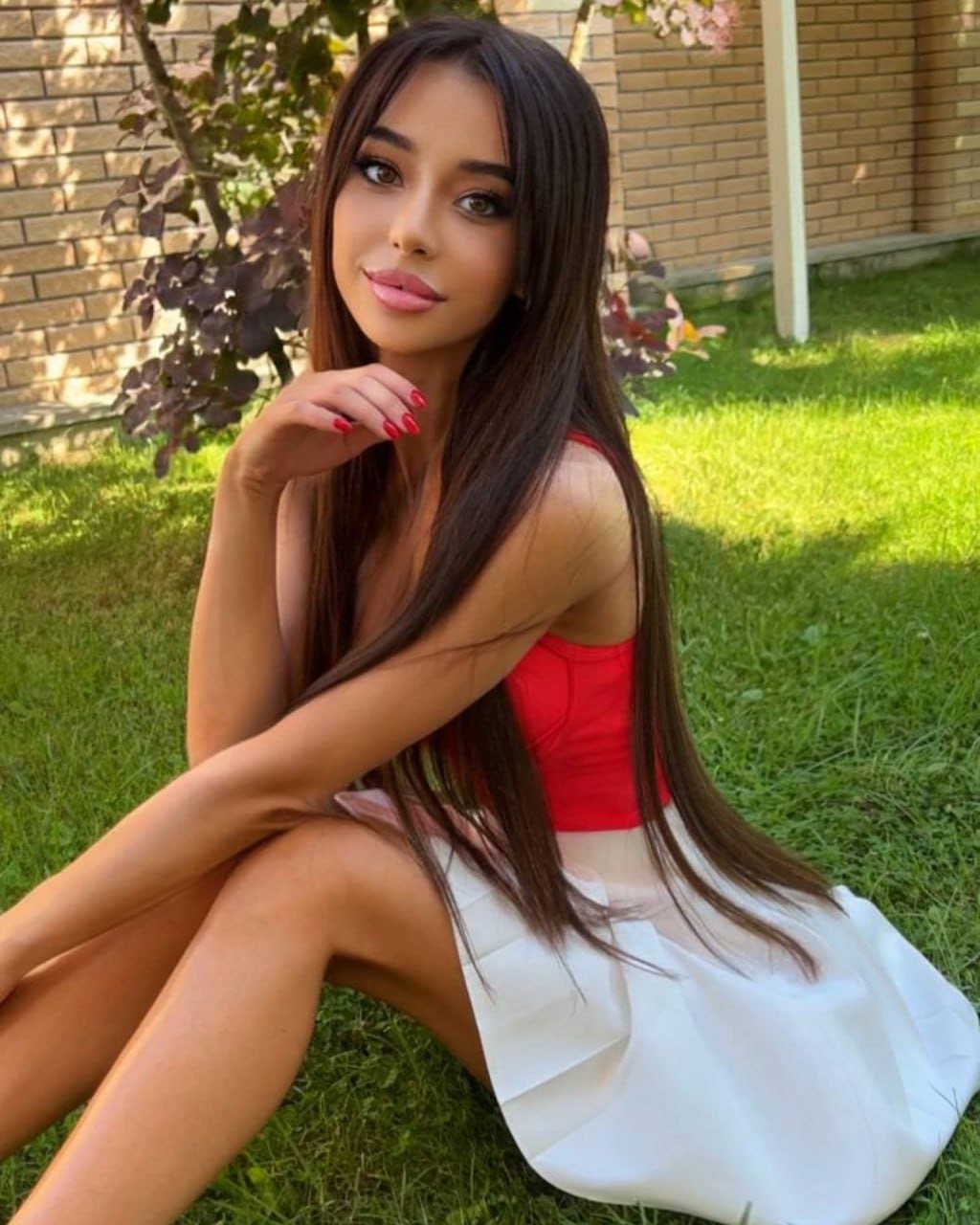 Beatrice sits on a lawn before a brick wall, wearing a red sleeveless top and white skirt, with one hand raised near her chin.