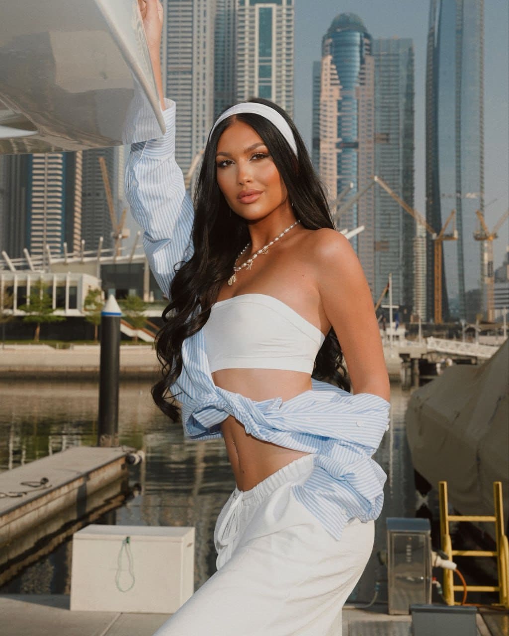 A woman stands on a dock before a city skyline, wearing a white crop top, striped shirt, and white pants, with one arm raised.