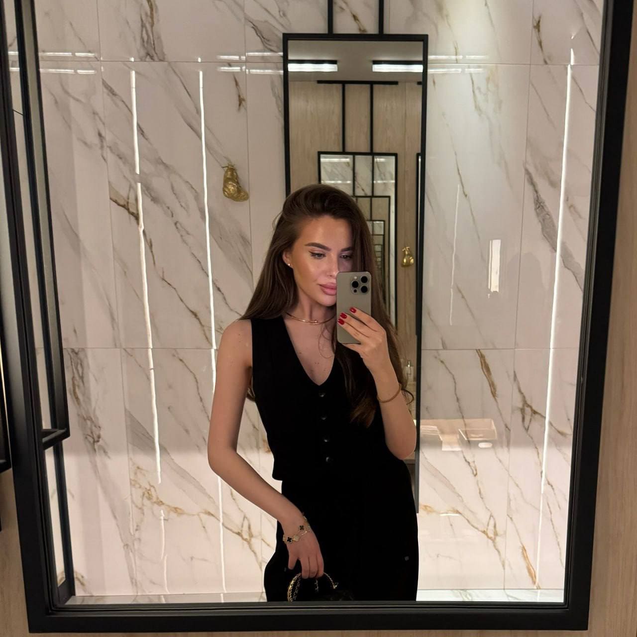 Mirror selfie in a marble-tiled room showing a woman in a sleeveless black button-front top, holding a smartphone and a small handbag.