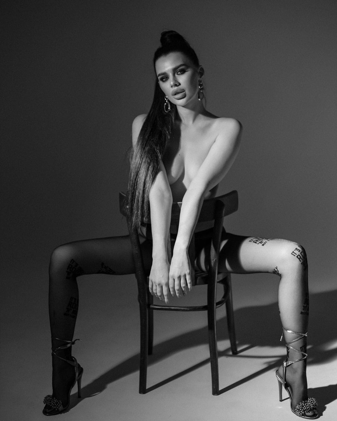 A woman with dark hair in a high bun sits on a wooden chair wearing sheer patterned tights and strappy heels, arms resting on the chair back in a studio.