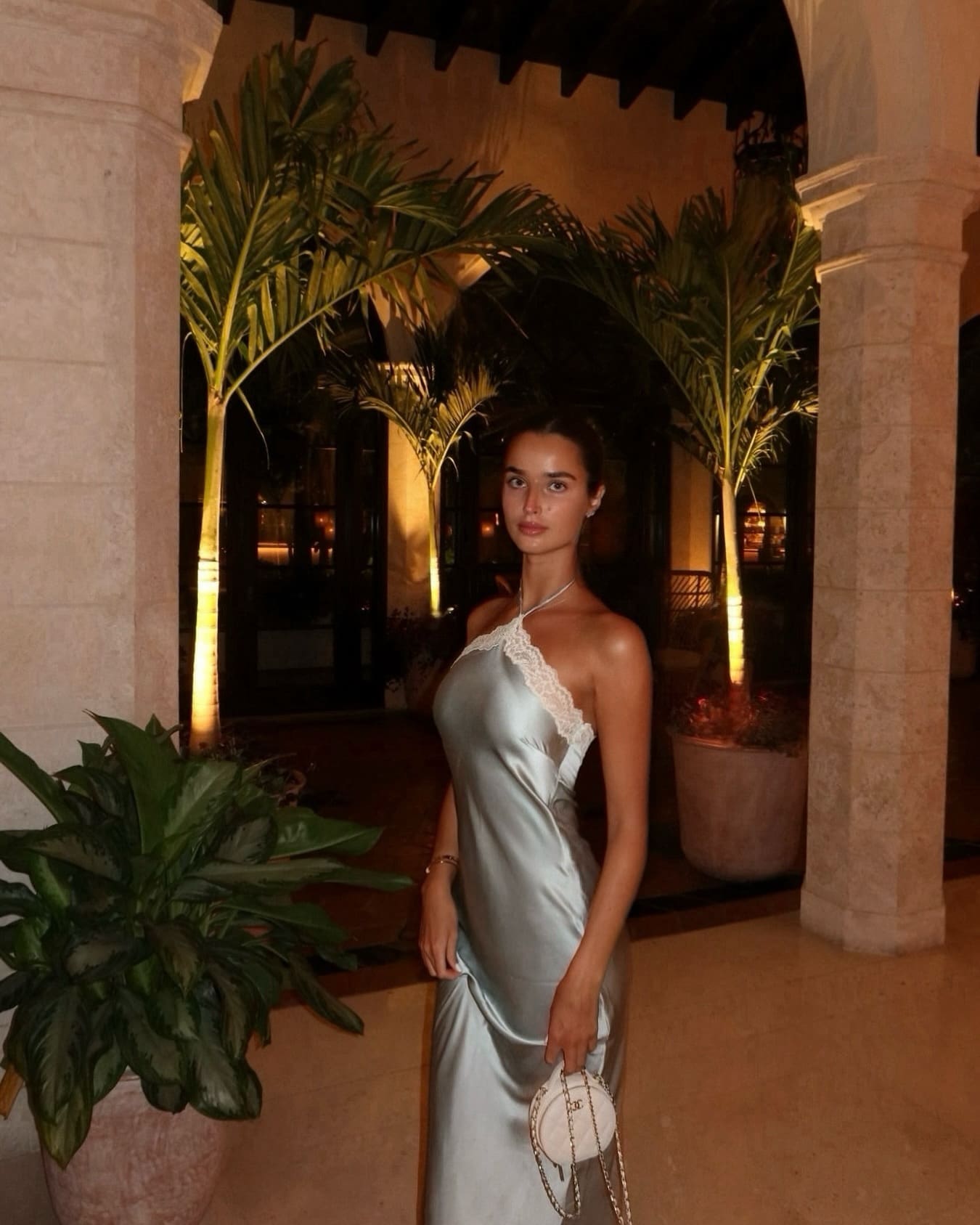 A woman in a light satin one-shoulder gown stands among stone columns and palms, holding a small chain-strapped handbag.