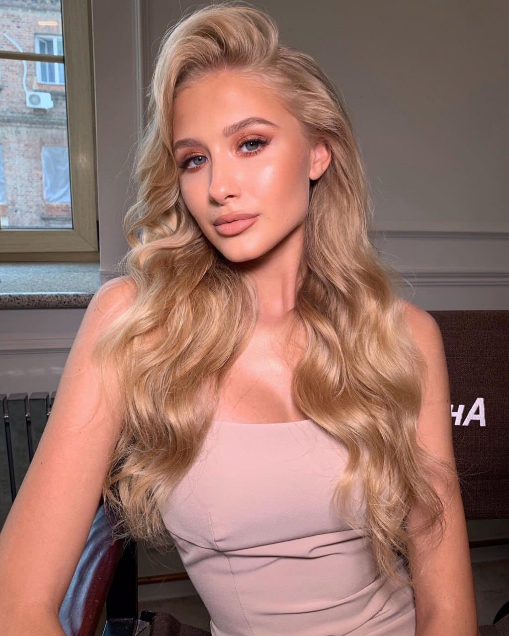Maria sits indoors before a window, wearing a sleeveless light pink top with a straight neckline and long wavy blonde hair.