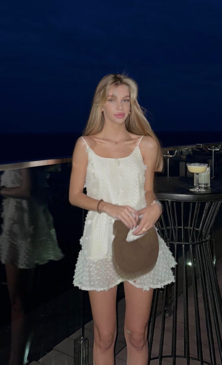 A woman in a short white scalloped dress holds a brown hat at an outdoor bar at night.