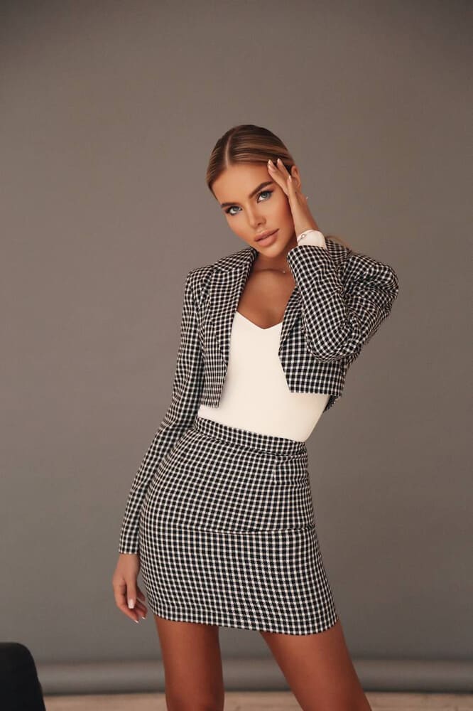 Woman with long brown hair stands in a studio wearing a black-and-white checkered cropped blazer and mini skirt over a white top, hand raised to head.