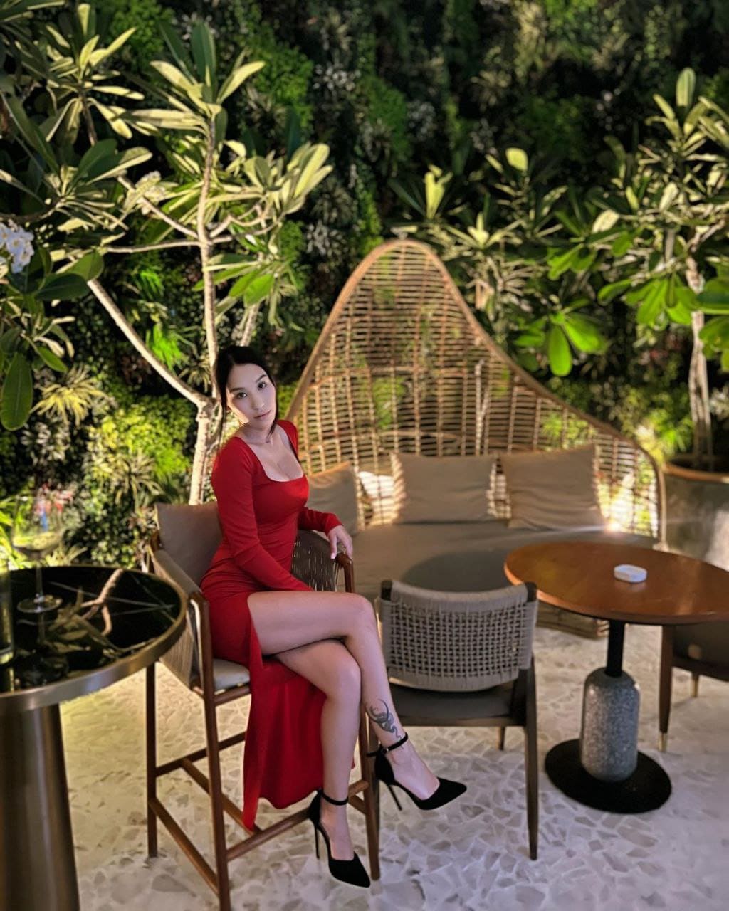 A woman sits on a wooden chair outdoors in a red dress with a high slit, surrounded by green foliage and a wicker sofa.