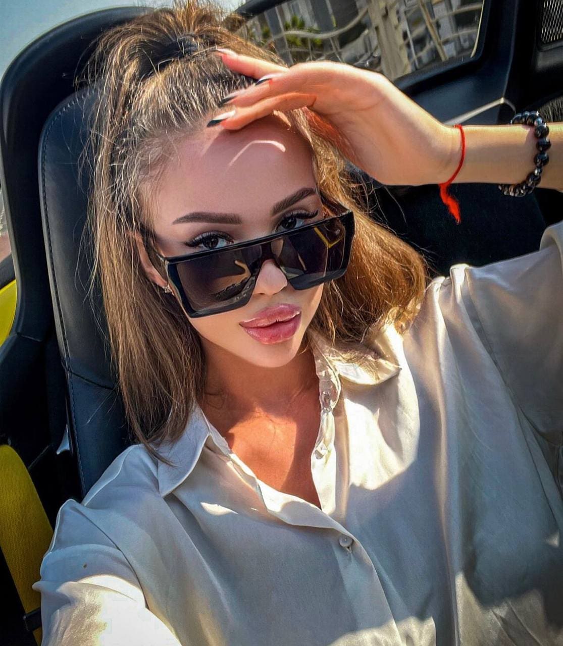 A woman with a high ponytail sits in a car wearing a loose white shirt and dark sunglasses, hand raised to her forehead.
