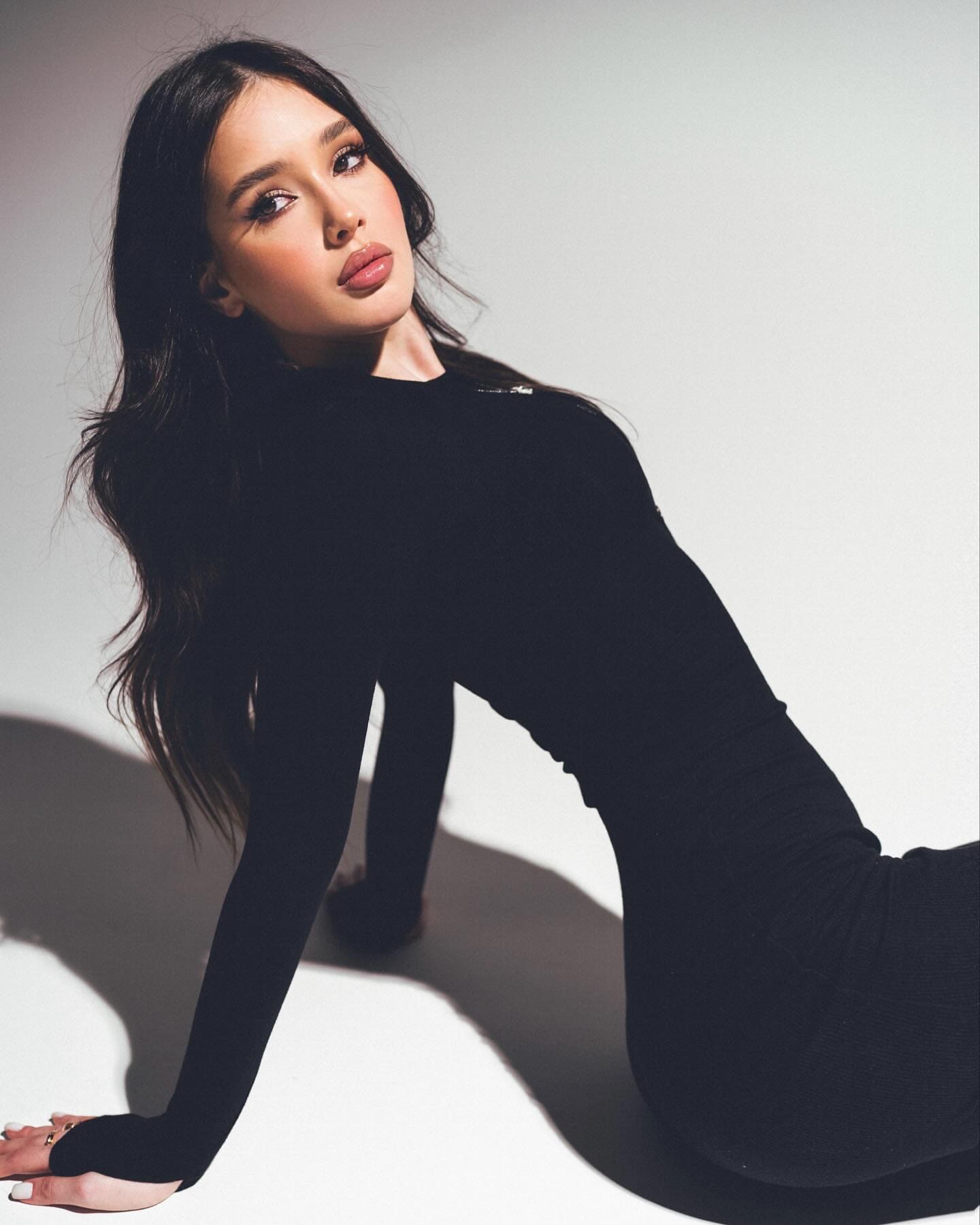 A woman in a black long-sleeved dress sits on a white floor, leaning back on one arm against a plain light background.
