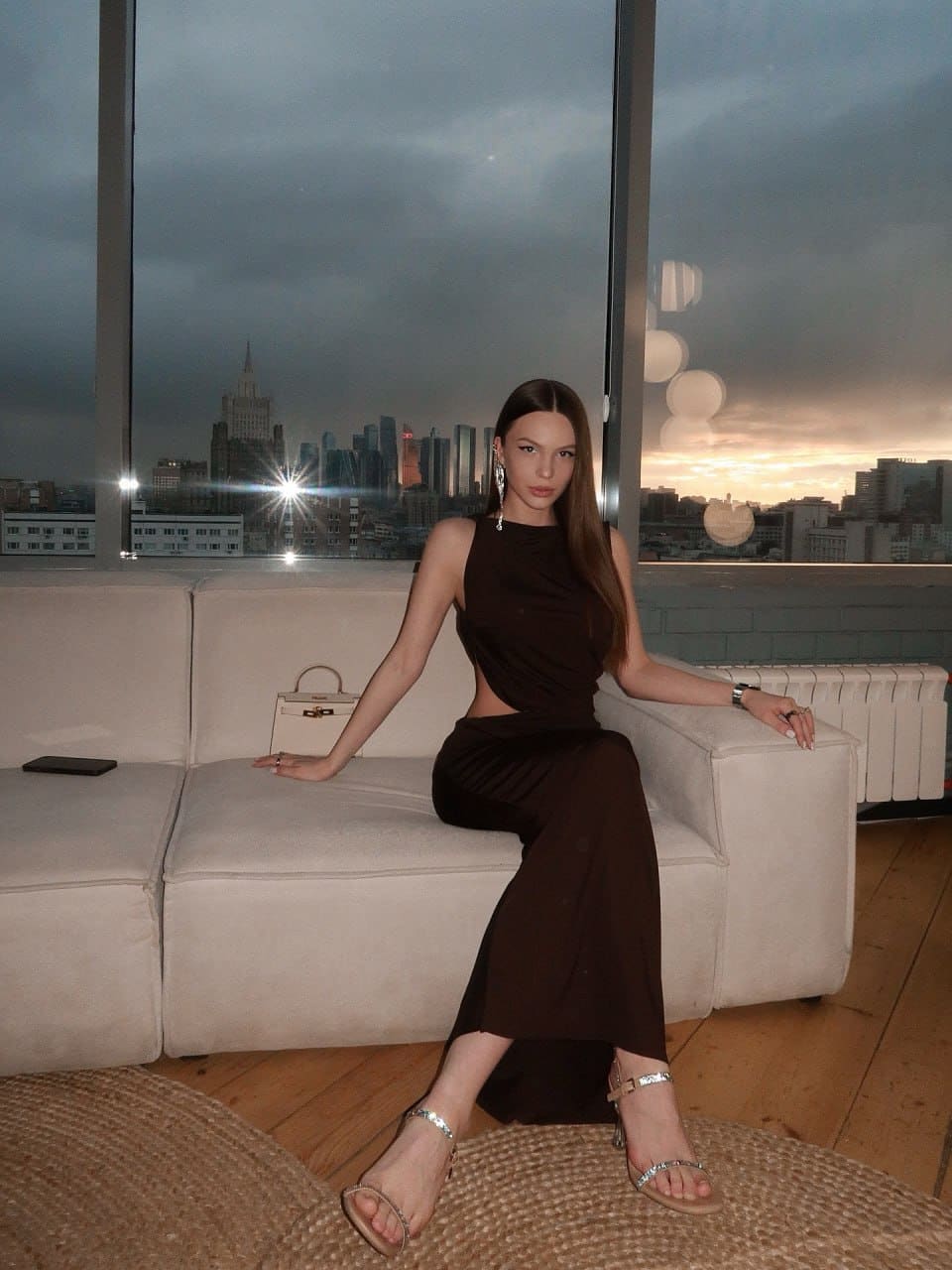 A woman sits on a white sofa before large windows showing a dusk city skyline, wearing a dark brown dress with a side cutout and strappy heels.