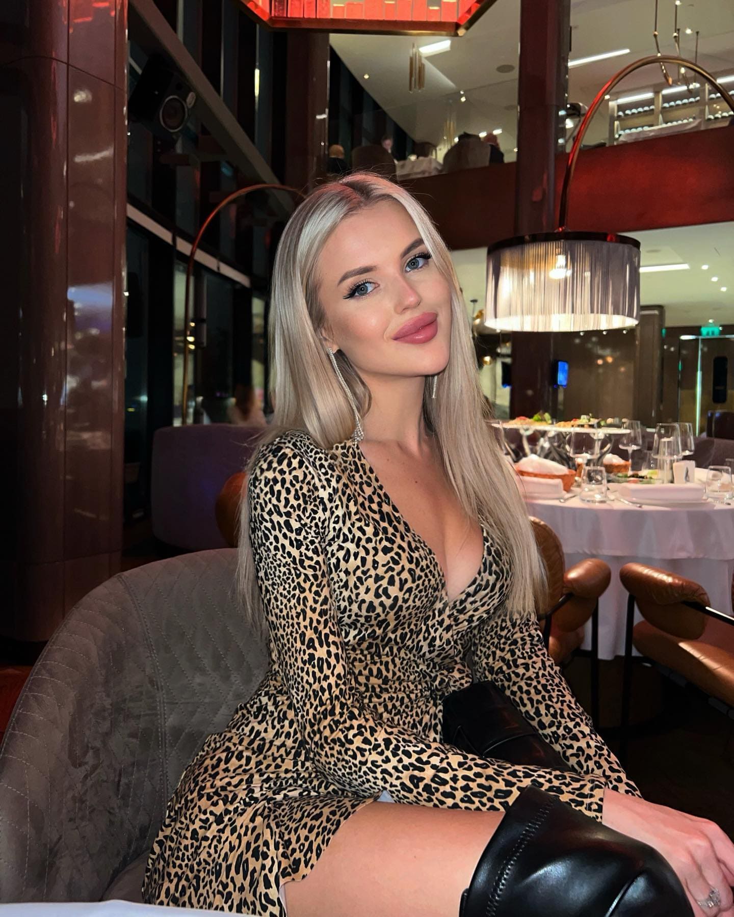 A woman in a leopard-print dress and black boots sits in a restaurant booth with a dining table and warm lighting behind her.