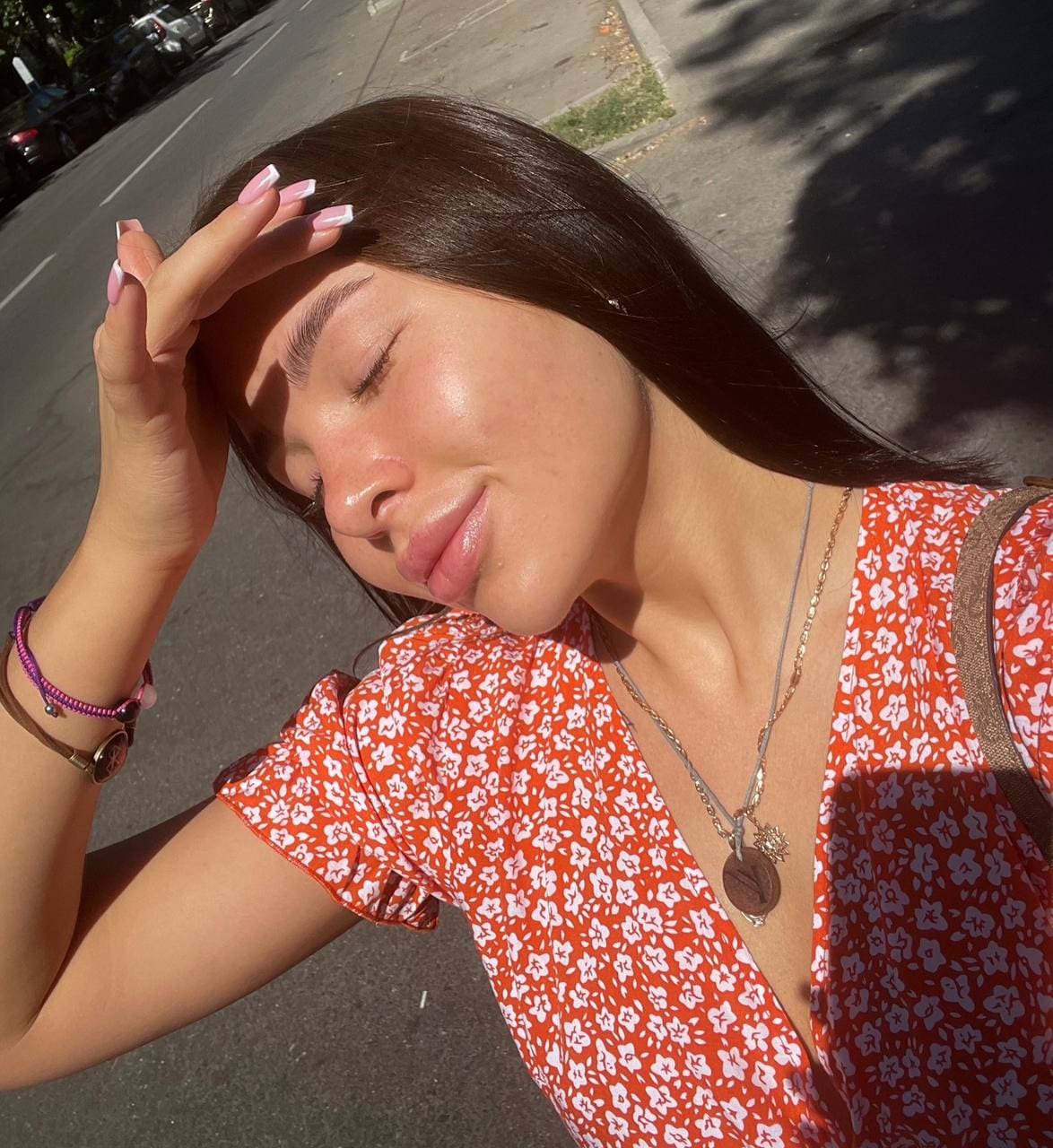 Woman with long dark hair and pink nails stands on a sunny street in a red floral top, hand to forehead with eyes closed.