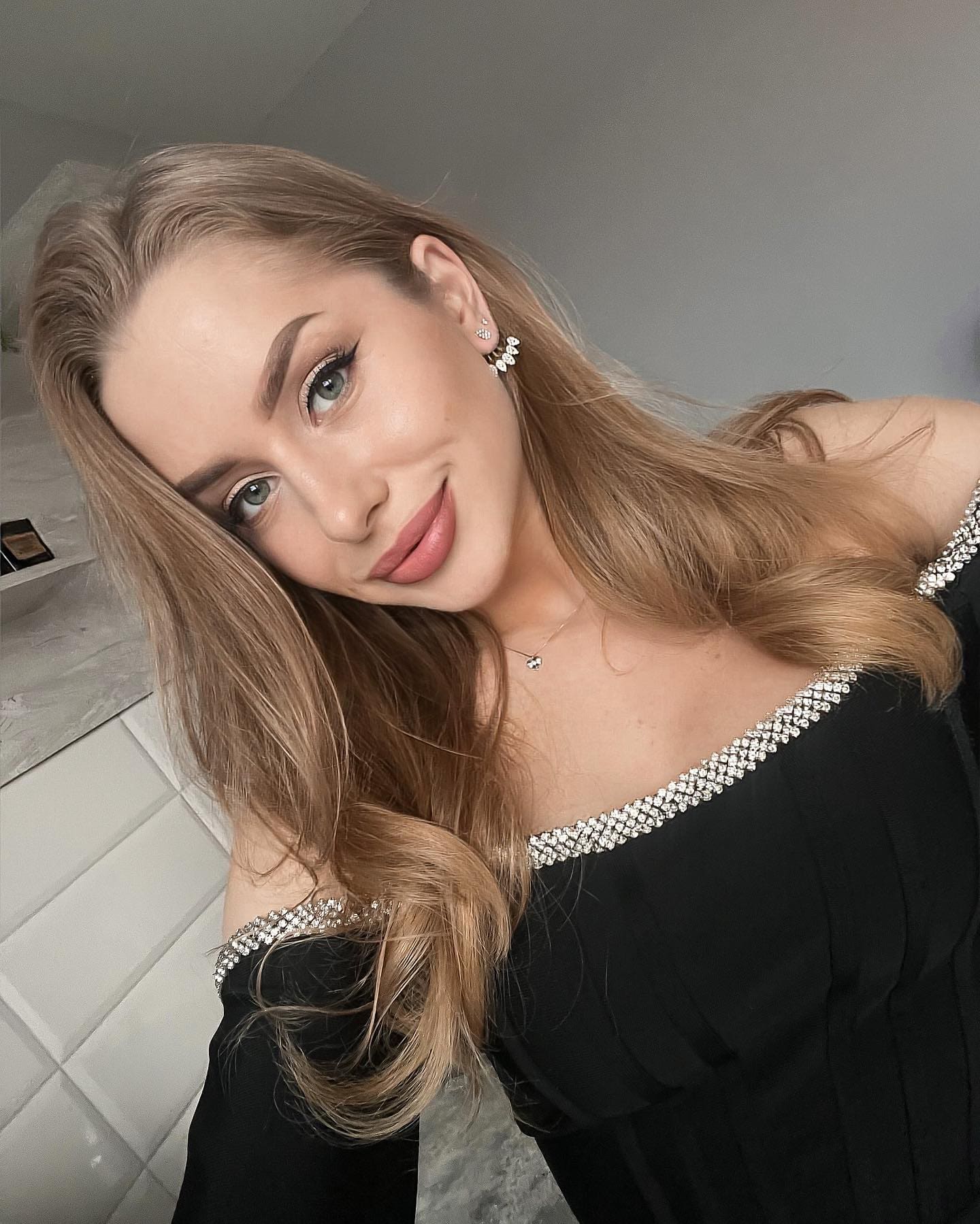 A woman tilts her head wearing a black off-the-shoulder top with rhinestone trim, in a room with a padded white wall.
