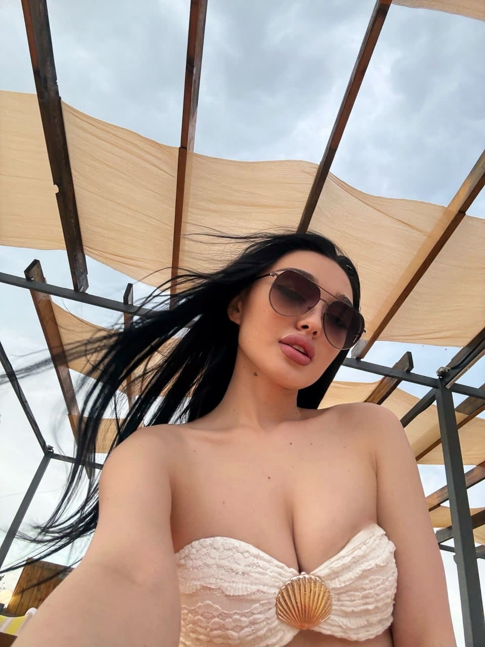 Low-angle selfie of a woman in sunglasses and a cream lace top, standing under a wooden pergola against a cloudy sky.