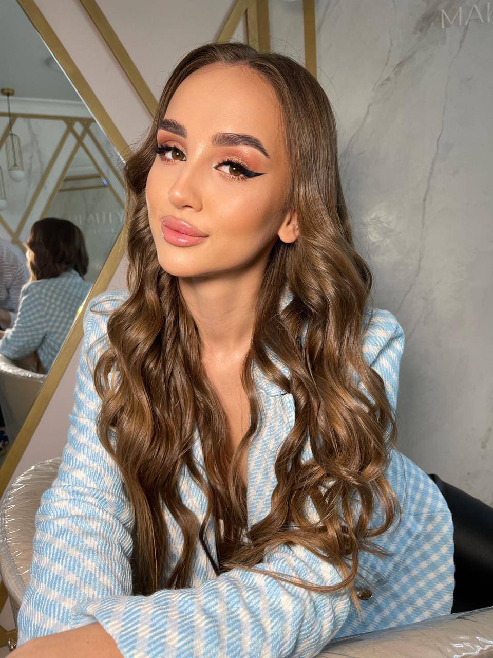 A woman sits in a chair wearing a light blue and white checkered blazer, positioned in front of a mirror and a marble wall.
