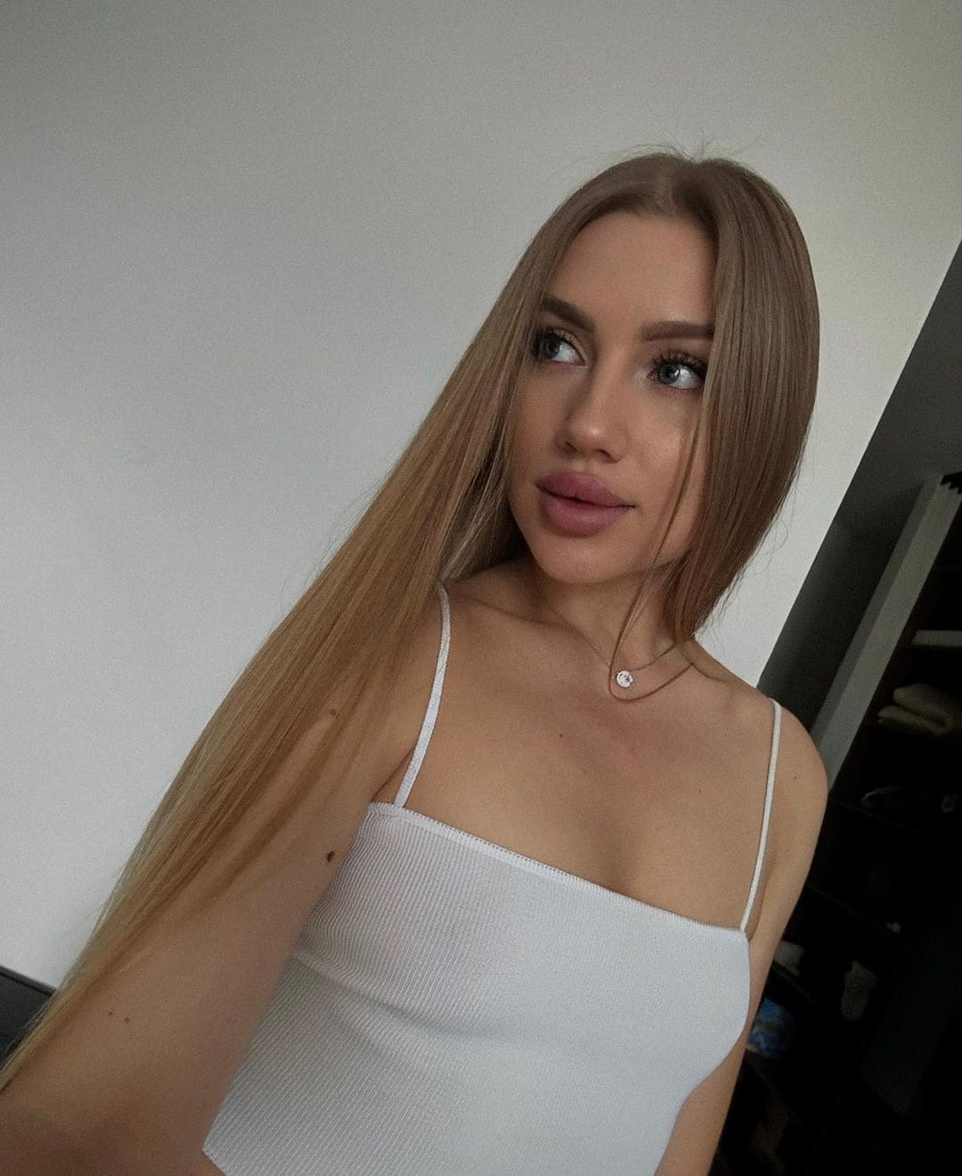 Woman with straight light-brown hair in a white ribbed top and necklace, looking sideways against a plain wall.