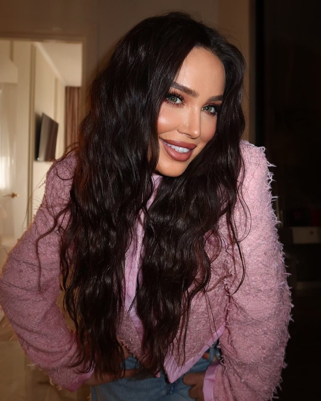 Indoors, she smiles wearing a textured pink cardigan over a light top and blue jeans, with a doorway in the background.