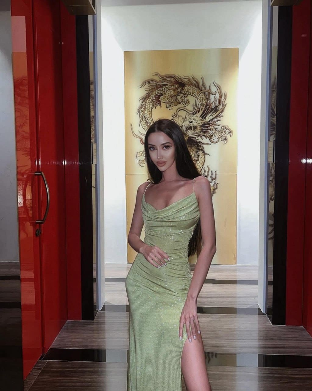 Linetta stands in a hallway with red doors and gold dragon art, wearing a sparkly light green gown with a high slit.