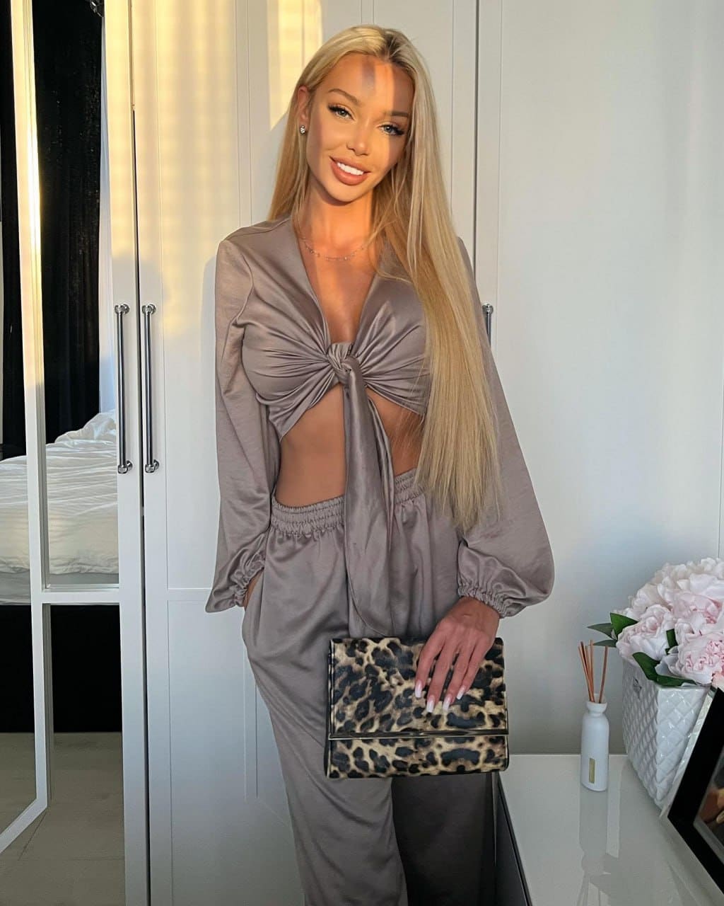 Naomi stands by white wardrobes in a taupe satin knotted top and trousers, holding a leopard-print clutch.