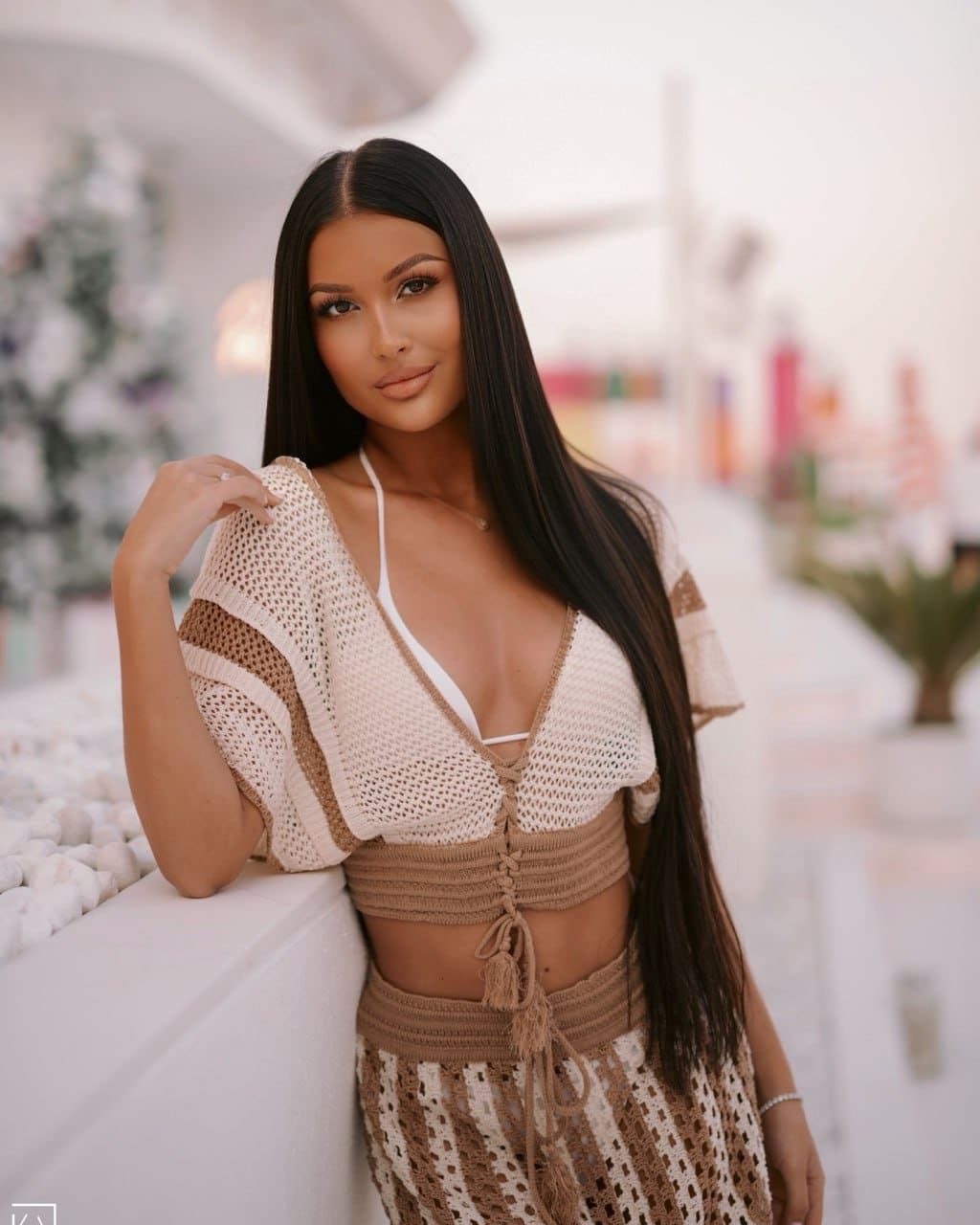 A woman leans on a white ledge indoors, wearing a beige and brown crochet two-piece outfit with a deep V-neckline.