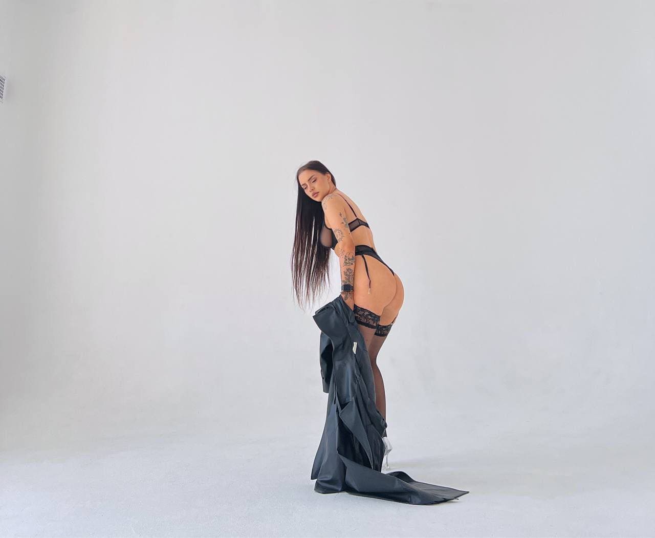 A tattooed woman in black lace lingerie and stockings stands in a white studio, holding dark fabric while looking back.