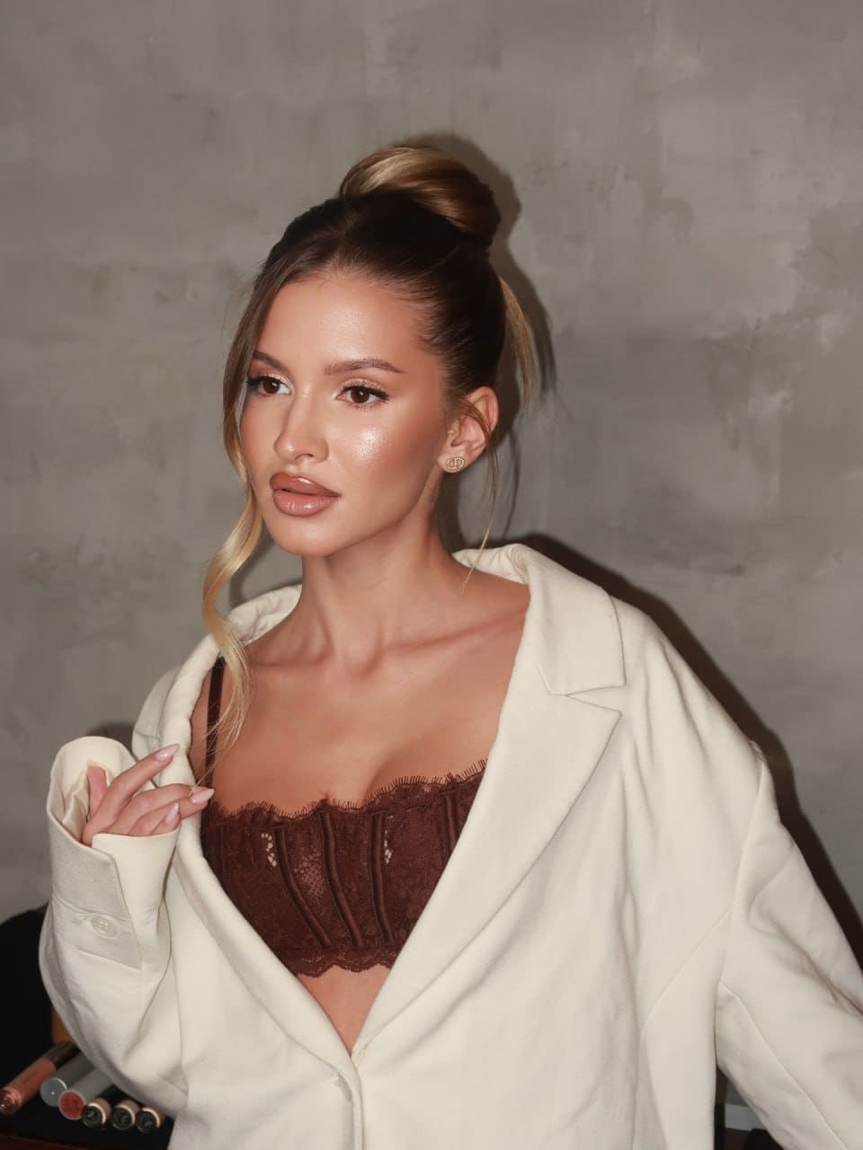 Luna stands against a gray wall in an open white blazer over a brown lace bustier, her blonde hair in a high bun.