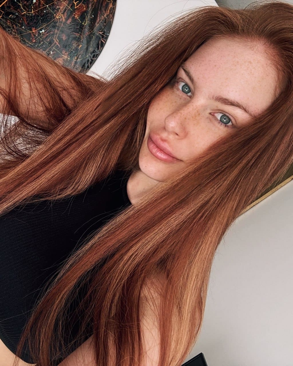 Close-up selfie of a freckled woman with auburn hair in a black top, hand behind head, against a white wall with a circular object.