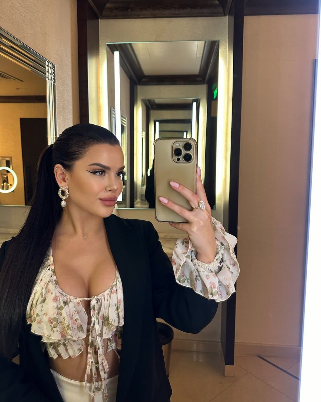 Sophia takes a mirror selfie in a hallway, wearing a black blazer over a floral ruffled crop top and white bottoms.