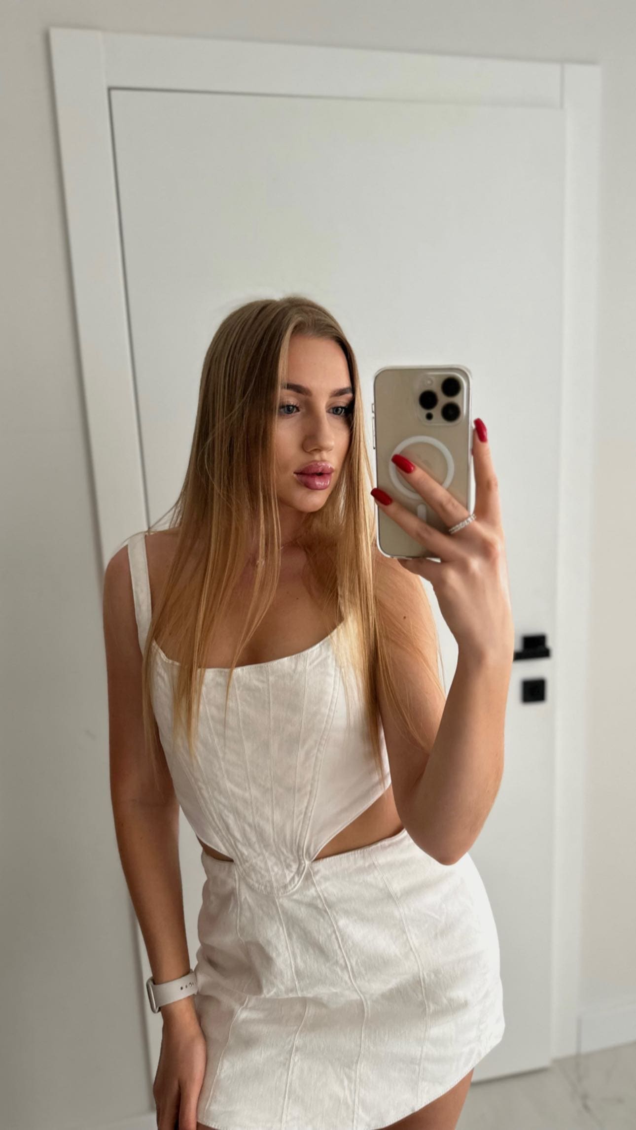 Mirror selfie of a woman in a white sleeveless top and short skirt, standing before a white door.