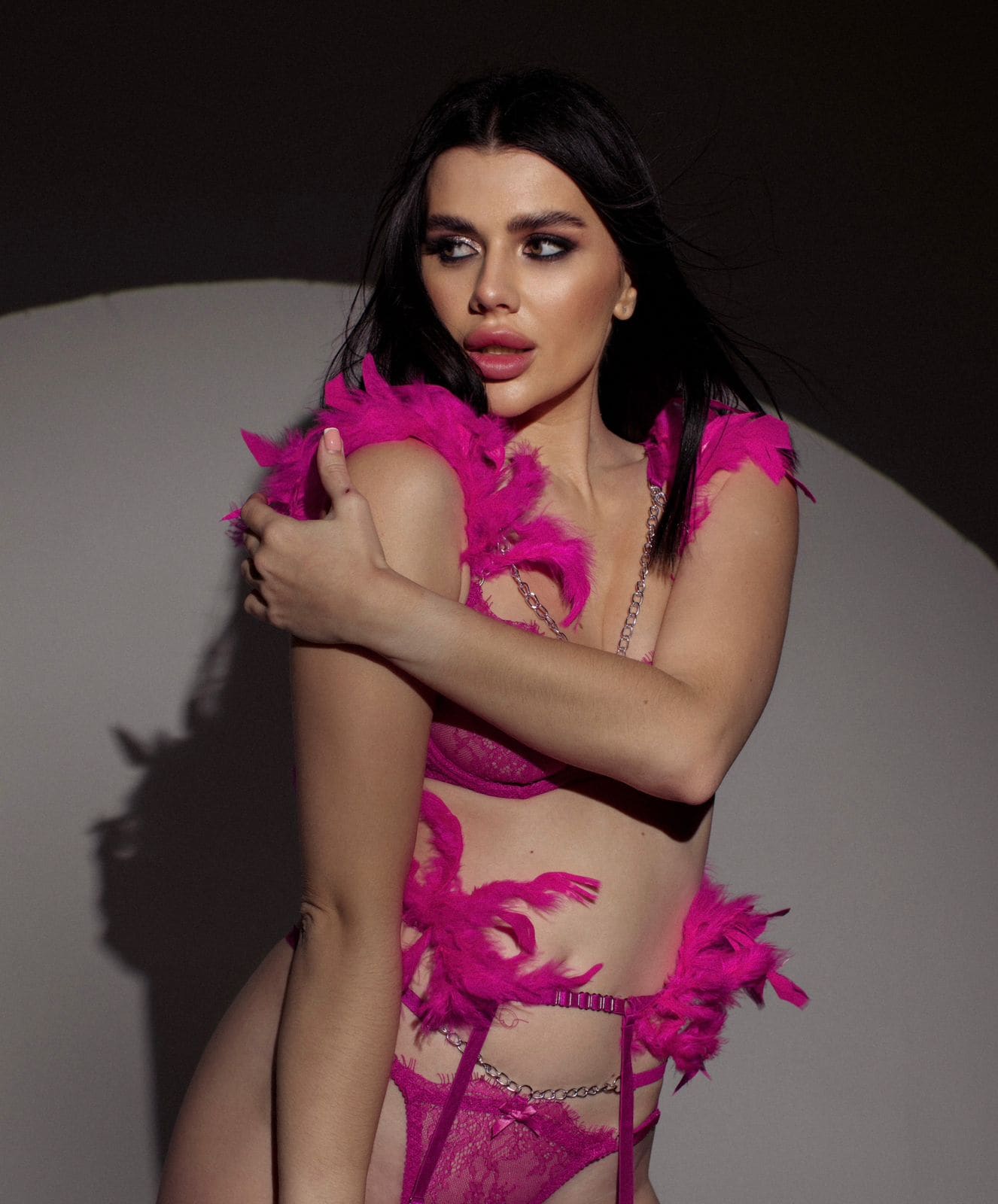 A woman with long dark hair stands against a gray backdrop in a bright pink feathered lace lingerie set with chain accents, arms crossed over her chest.