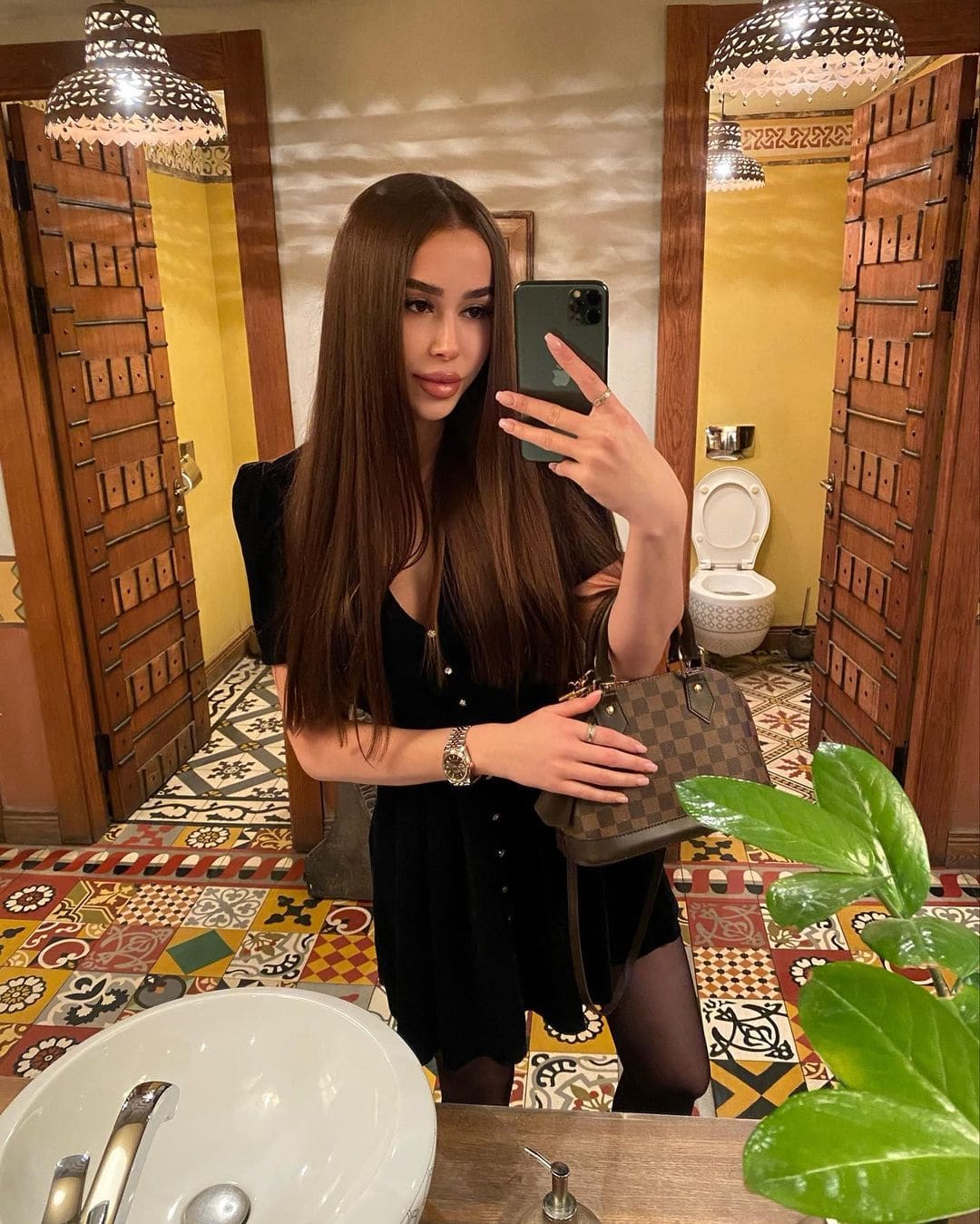 A woman in a black short-sleeved dress holds a patterned bag while taking a mirror selfie in a bathroom.