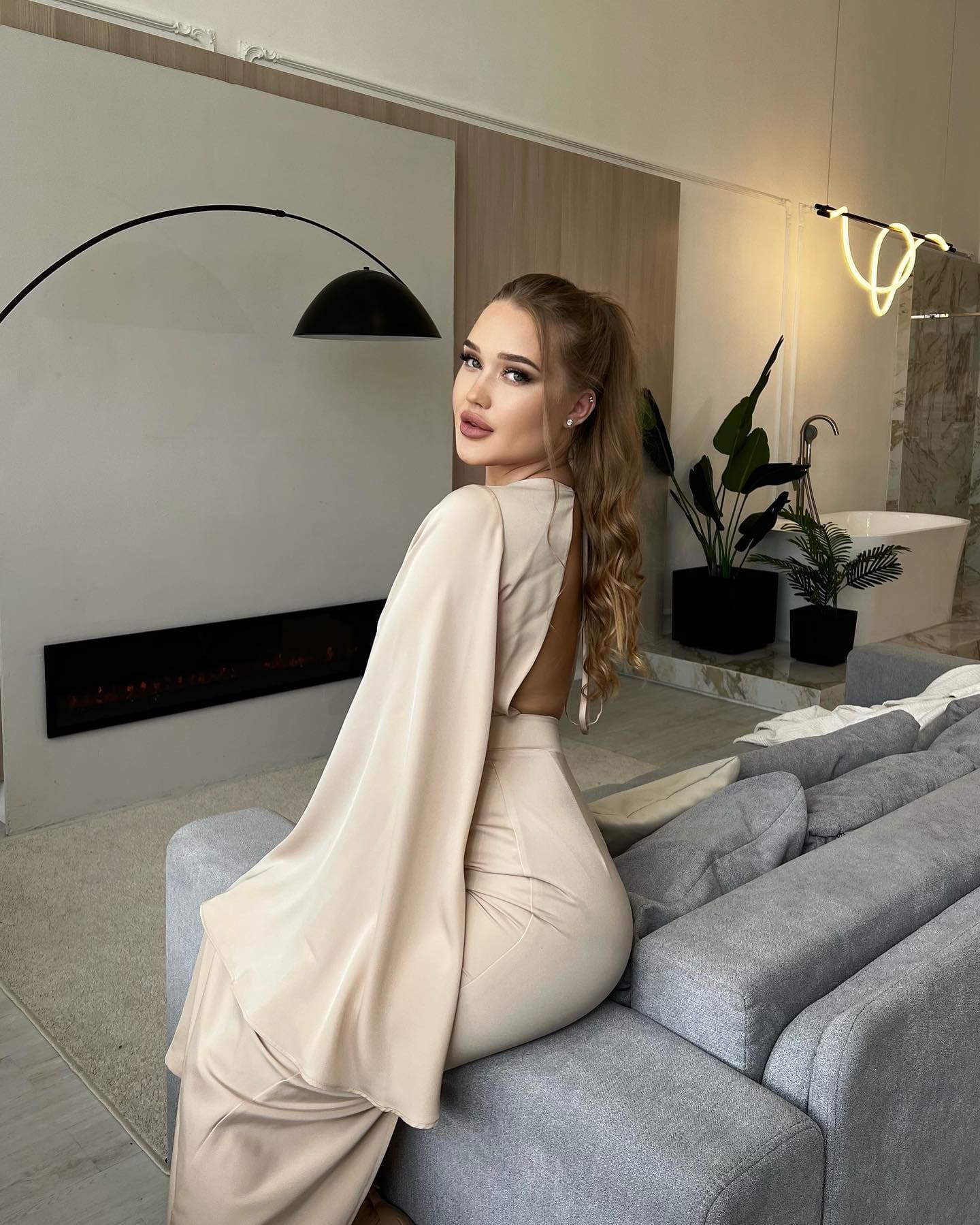 Woman with a high ponytail sits on a grey sofa in a beige satin outfit, looking over her shoulder in a modern room.