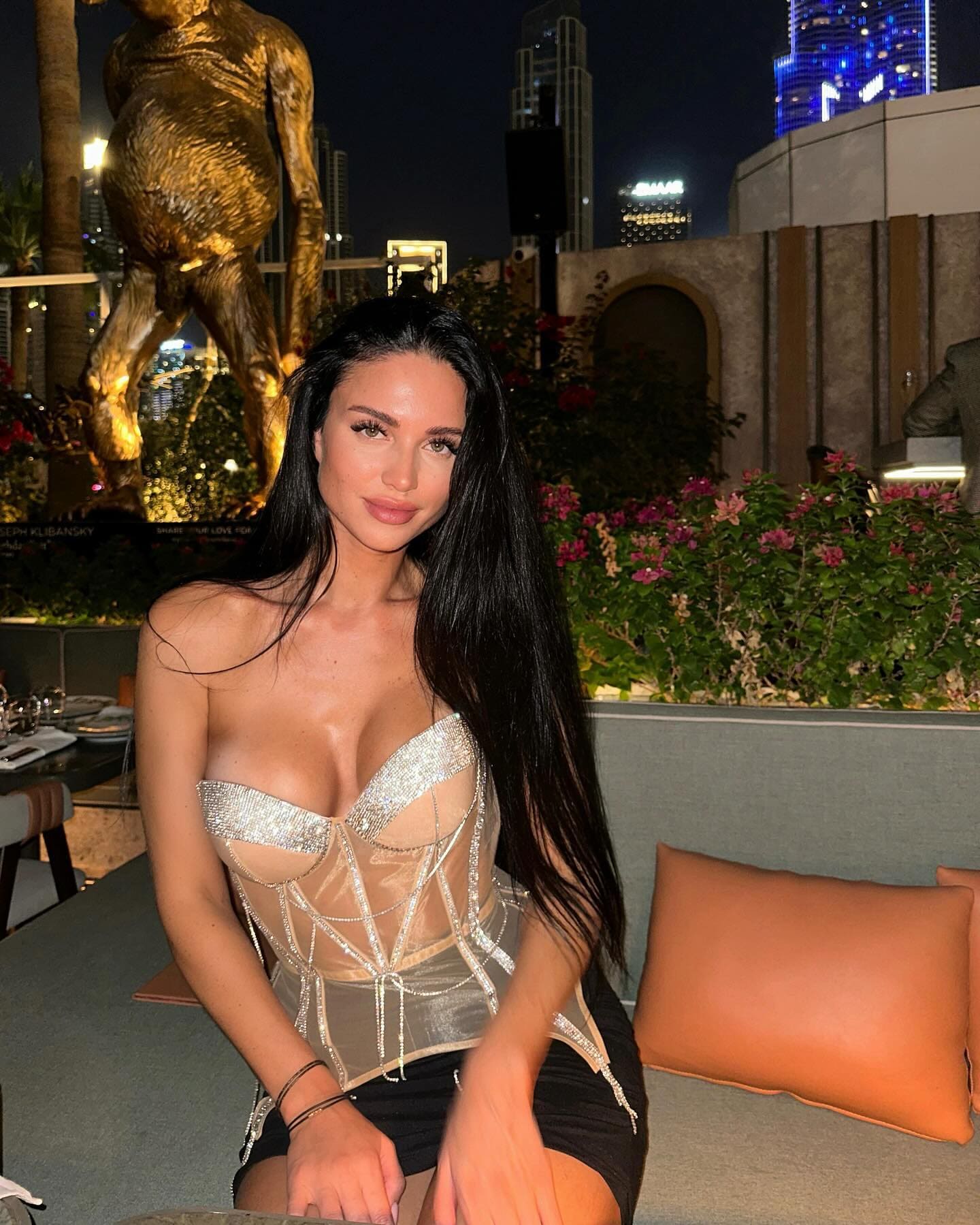 A woman in a sheer beaded corset and black skirt sits on a night lounge, with a golden statue and city lights behind her.