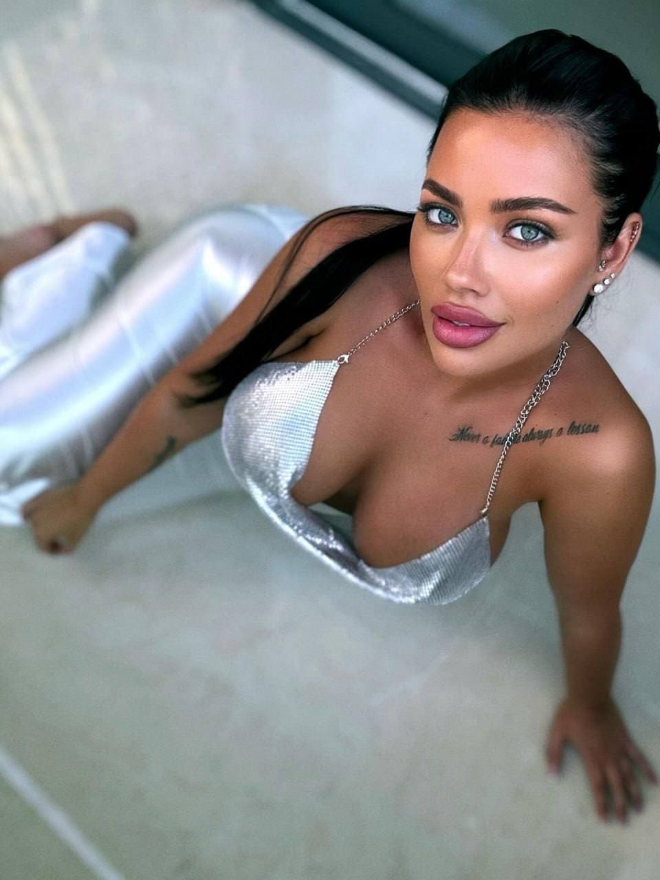 A woman lies on a light floor in a silver sequined mini dress with chain straps, looking up at the camera.