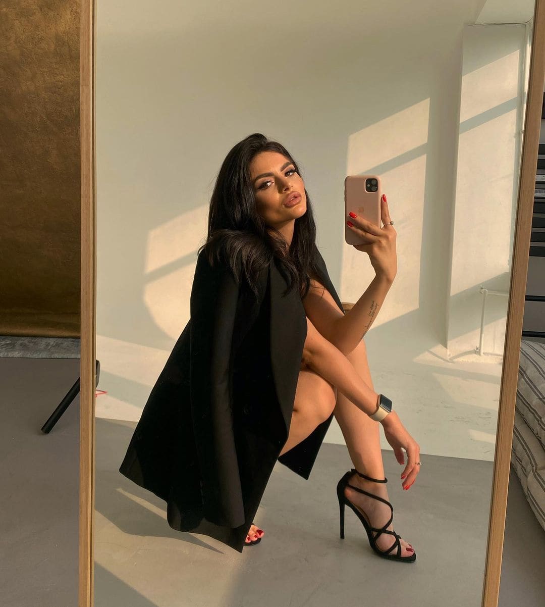 A woman takes a mirror selfie in a black open coat and strappy heels, holding a phone in a sunlit room.