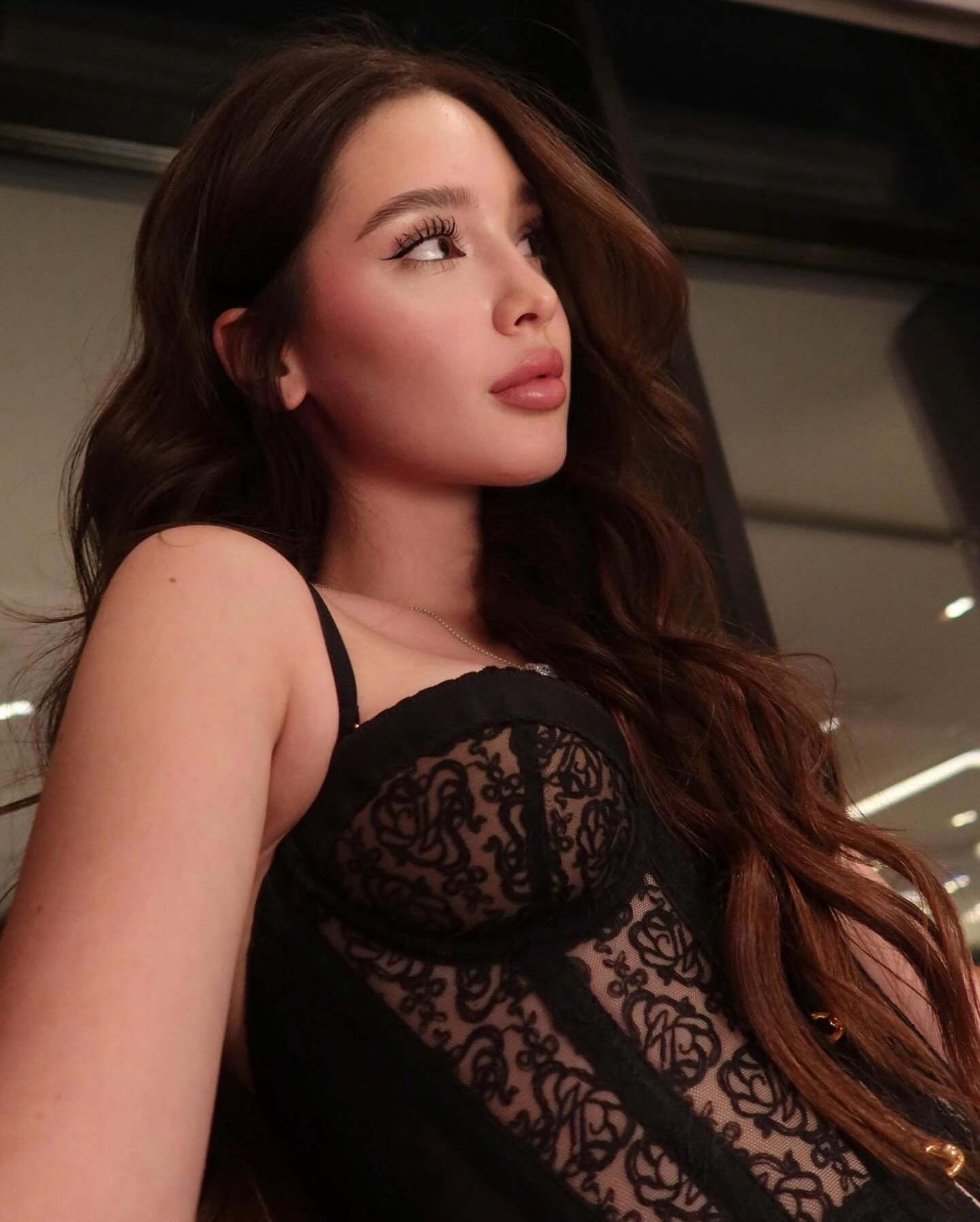 A chest-up view of a woman in a black lace top with sheer panels, looking upward against a blurred indoor background.