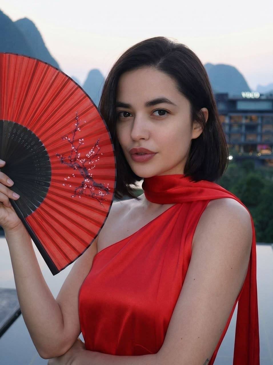 Lilian holds a red floral fan in a red satin wrap dress, standing outdoors against mountains and a building at dusk.