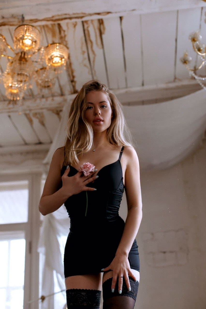 A blonde woman in a black slip dress and lace stockings holds a pink rose to her chest under a chandelier and distressed ceiling.