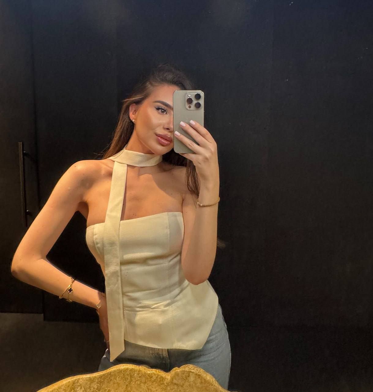 A mirror selfie in a dark room features a woman in a cream halter-neck top with a long tie and light blue jeans, holding a grey smartphone.