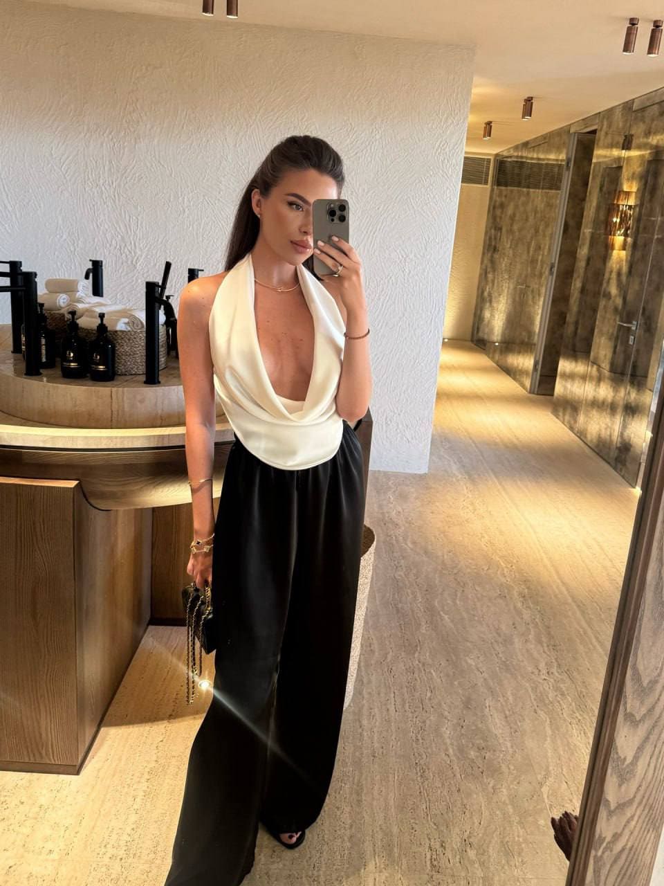 In a modern bathroom, a woman with a ponytail takes a mirror selfie wearing a white halter top and black wide-leg trousers, holding a black handbag.