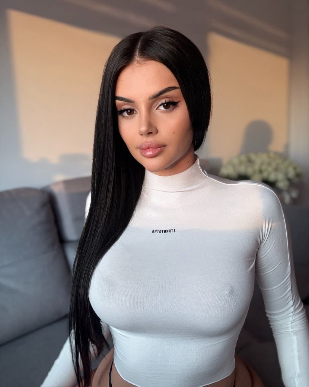 Valentina stands indoors in a white turtleneck, facing the camera near a gray sofa and vase of white flowers.