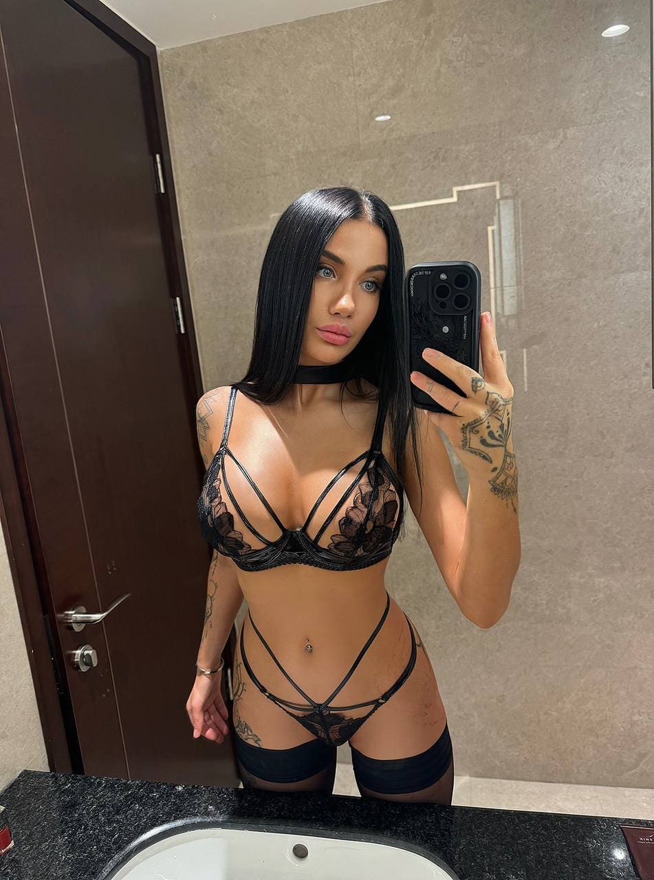 Mirror selfie in a bathroom showing a woman in black lace lingerie with strappy details and thigh-high stockings.